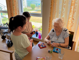 Zwei Kinder stehen an einem Tisch und spielen mit einer älteren Dame. Auf dem Tisch liegen verschiedene Brettspiele und einige bunten Spielfiguren. Im Hintergrund sind Fenster mit Vorhängen und Pflanzen sichtbar.