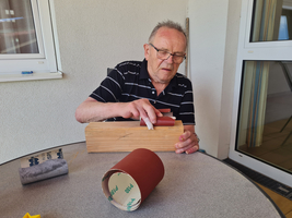 Ein älterer Mann bearbeitet Holz mit Schleifpapier auf einem Tisch.