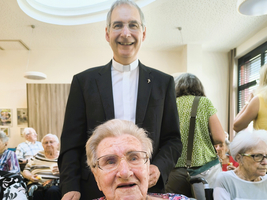 Ein lächelnder Priester steht hinter einer älteren Frau im Seniorenheim.
