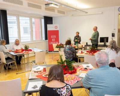 Aufnahme von Adventfeier für Senioren, Senioren von hinten fotografiert, sitzen an weihnachtlich dekorierten Tischen, vorne zwei Personen die stehen und erzählen 
