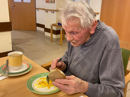 Ein älterer Mann isst ein Brötchen mit Eiern am Tisch.
