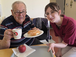 Älterer Mann mit Brille sitzt an einem Tisch und lächelt, während er eine Tasse hält. Vor ihm befindet sich ein Teller mit einem Brötchen und ein Apfel auf einem Tablett. Eine junge Frau in einem rot-braunen Oberteil steht neben ihm und lächelt ebenfalls.