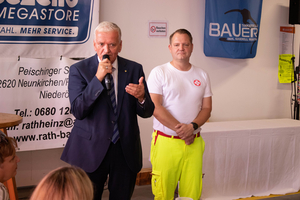 Ein Mann spricht am Mikrofon, während ein anderer in hellen Arbeitskleidern zuhört.