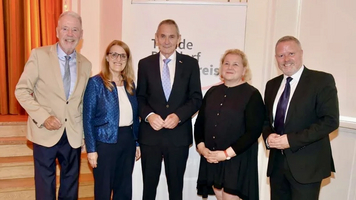 Gruppenshot von fünf Personen, die zusammen stehen und lächeln. Sie tragen formelle Kleidung und posieren vor einem Banner mit dem Schriftzug 