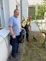 Zwei Männer stehen im Hof eines Hauses und arbeiten im Garten. Einer hält einen schwarzen Müllsack, während der andere mit einem Rechen arbeitet. Um sie herum sind einige Pflanzen und das Gebäude ist sichtbar.