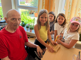 Ein älterer Mann sitzt an einem Tisch und schaut freundlich in die Kamera, während mehrere Kinder um ihn herumstehen und an einem kleinen Paket arbeiten. Der Raum hat große Fenster mit Blick ins Grüne und ist hell erleuchtet.