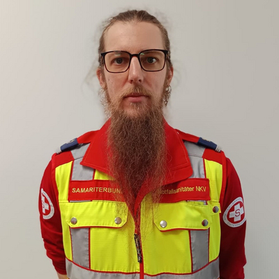 Ein Mann mit Brille und einem langen Bart steht vor einem neutralen Hintergrund. Er trägt eine leuchtend gelbe Weste mit roten Akzenten, die das Logo einer Rettungsorganisation zeigt. Der Mann sieht ernst aus und steht aufrecht.