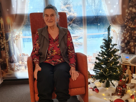 Eine ältere Frau sitzt in einem Sessel vor einem weihnachtlichen Hintergrund.