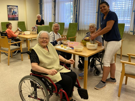 Eine Gruppe älterer Frauen und eine Betreuerin beim gemeinsamen Essen in einem Raum.
