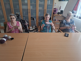 Drei ältere Frauen sitzen an einem Tisch und stricken. Jede hält Wolle und Stricknadeln in den Händen. Im Hintergrund sind ein Rollstuhl und Regale mit verschiedenen Gegenständen zu sehen. Die Atmosphäre wirkt freundlich und entspannt.