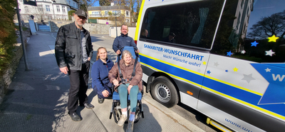 Eine ältere Frau sitzt in einem Rollstuhl und wird von drei Personen umgeben. Sie stehen an einem Fahrzeu g für die „Samariter-Wunschfahrt“. Die Szene ist sonnig, mit einem Gebäude im Hintergrund. Alle wirken freundlich und hilfsbereit.