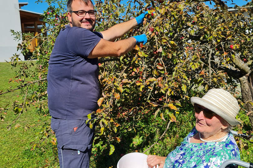 Ein Mann pflückt gemeinsam mit einer älteren Frau, die im Rollstuhl sitzt, Äpfel von einem Baum. Die Frau hält einen Eimer, während der Mann frisches Obst erntet. Im Hintergrund sind ein blauer Himmel und ein Garten sichtbar.