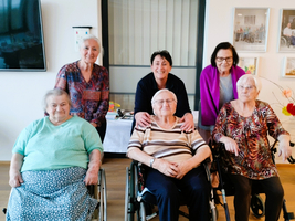 Eine Gruppe von sechs Frauen, darunter eine Betreuerin, die in einem hellen Raum zusammensteht. Vier Frauen sitzen in Rollstühlen, während zwei hinter ihnen stehen. Alle lächeln und scheinen sich wohlzufühlen.