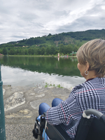 Frau im Rollstuhl blickt auf einen ruhigen See mit grüner Umgebung.