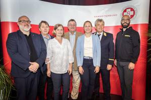 Sechs Personen stehen in einer Reihe vor einer roten Hintergrundwand des Samariterbundes.