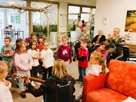 Eine Gruppe von Kindern singt und tanzt in einem Gemeinschaftsraum. Im Vordergrund sitzt eine Person mit einer Gitarre. Ältere Menschen schauen von ihren Plätzen aus zu. Der Raum ist freundlich und hell gestaltet.