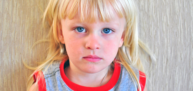 Ein kleines Kind mit blonden Haaren und blauen Augen schaut ernst in die Kamera.