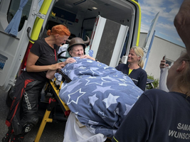 Eine ältere Person wird auf einer Trage aus einem Rettungswagen transportiert. Um sie herum stehen mehrere Rettungskräfte, die freundlich zu ihr sprechen. Die Person ist mit einer Decke mit Sternenmustern bedeckt. Im Hintergrund sind Bäume und ein blauer Himmel zu sehen.