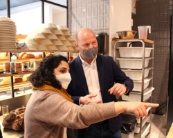 Peter Ostendorf von mel&koffie, Sya Ahmad, Bezirksvorsteherin neunter Bezirk in mel&koffie Filiale im neunten Bezirk, zeigen auf etwas, dass im Bild nicht mehr ersichtlich ist, im Hintergrund Brot in Regal