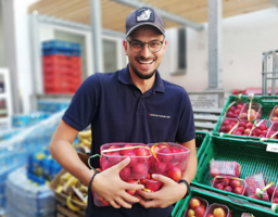 Mann mit blauem Samariterbund Wien Polo und blauer Kappe, Brille, Bart, hält Obst in Hand, im Hintergrund Sozialmarkt mit Obst und Gemüse in Kisten