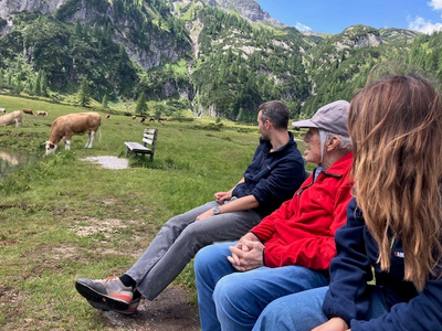 Drei Personen sitzen auf einer Bank und beobachten Kühe in der Landschaft.