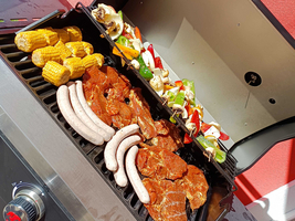 Auf einem Grill liegen verschiedene Grillwaren: mariniertes Fleisch, Würstchen, Gemüse-Spieße und Maiskolben. Der Grill ist geöffnet und sieht aus, als wäre das Grillen gerade im Gange. Im Hintergrund ist eine grüne Wiese und eine rote Wand sichtbar.