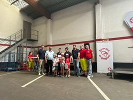 Eine Gruppe von Erwachsenen und Kindern steht in einer großen Lagerhalle zusammen. Sie lächeln und halten ein Geschenk. Im Hintergrund sind Regale und eine Treppe sichtbar. An der Wand hängen Plakate mit dem Schriftzug 