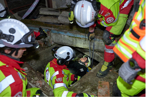 Eine Gruppe von Rettungskräften in leuchtend grünen und roten Uniformen arbeitet an einer Rettungsaktion. Sie helfen einer Person, die in einer tiefen Öffnung sitzt. Einige Rettungskräfte sind dabei, mit Ausrüstung zu manövrieren, während andere die Situation beobachten.