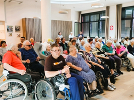 Eine Gruppe älterer Menschen sitzt in einem Gemeinschaftsraum. Einige verwenden Rollstühle. Die Atmosphäre ist lebhaft, mit mehreren Personen, die miteinander sprechen. Der Raum hat große Fenster, die viel Licht hereinlassen.