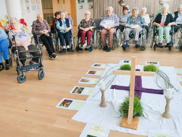 Eine Gruppe älterer Menschen sitzt in Rollstühlen oder auf Stühlen in einem Gemeinschaftsraum. In der Mitte des Raumes befindet sich ein mit einem weißen Tuch bedeckter Tisch, auf dem ein Holzkreuz, Kerzen und grüne Pflanzen stehen. Um das Kreuz sind mehrere Bilder und Beschreibungen angeordnet.