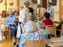 Eine Gruppe von älteren Menschen sitzt in einem hellen Raum. Einige Personen stehen oder bewegen sich. Es gibt mehrere Stühle und Tische. Im Hintergrund sind dekorative Elemente wie Schmetterlingsbilder und eine Uhr zu sehen.