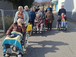 Eine Gruppe älterer Menschen in Rollstühlen und zu Fuß, die auf einem Gehweg spazieren. Einige tragen leichte Jacken, und die Sonne scheint. Im Hintergrund sind Bäume und ein Gebäude sichtbar.