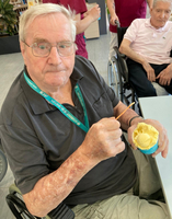 Ein älterer Mann mit Brille sitzt in einem Rollstuhl und hält eine Schüssel mit Eiscreme in der Hand. Er hat einen neugierigen Gesichtsausdruck und benutzt einen Spoon, um das Eis zu essen. Im Hintergrund sind weitere Personen sichtbar, die in einem Pflegeheim sind.