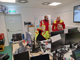 Drei Menschen in Warnkleidung stehen in einem modernen Büro mit mehreren Computermonitoren. Sie scheinen eine Besprechung zu haben und schauen auf einen der Bildschirme. Im Hintergrund sind technische Geräte und Kabel sichtbar.