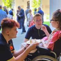 Eine Frau in einem Rollstuhl wird von zwei Personen beim Essen unterstützt.