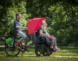 Rikschafahrt im Grünen, eine Person tritt in Pedale lächelt, zwei Personen sitzen in Rikscha und lächeln, Helm hängt auf Fahrradhalterung