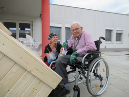 Frau und Mann im Rollstuhl bemalen gemeinsam ein Holzobjekt im Freien.