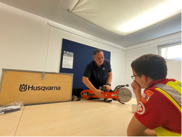 Ein Mann in einem blauem Hemd zeigt einem jungen Mann in einer Sicherheitsweste eine Handsäge auf einem Tisch. Im Hintergrund steht eine Holzkiste mit dem Logo von Husqvarna. Der junge Mann beobachtet aufmerksam.