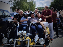 Eine Gruppe von Menschen sitzt zusammen auf einem gelben Motorrad mit Beiwagen. Sie lächeln und posieren fröhlich für die Kamera, umgeben von anderen Fahrzeugen und einer grünen Umgebung. Der Himmel ist teilweise bewölkt.