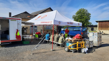 Rettungskräfte sitzen unter einem Zelt, im Hintergrund ein Einsatzfahrzeug und Ausrüstung.