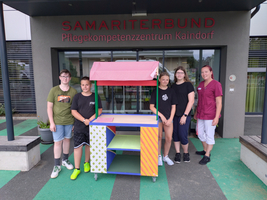 Eine Gruppe von fünf Personen steht vor einem bunten Verkaufsstand.