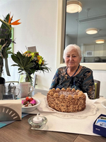 Eine ältere Frau sitzt mit einer Torte und Blumen am Tisch.