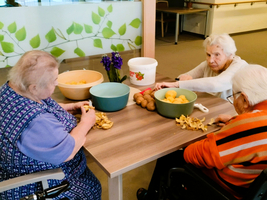 Drei ältere Frauen schälen Kartoffeln an einem Tisch in einem Gemeinschaftsraum.