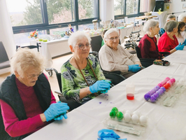 Ältere Frauen, die in einem hellen Raum sitzen und Ostereier bemalen. Sie tragen Handschuhe und konzentrieren sich auf ihre Arbeiten. Auf dem Tisch sind gefärbte Eier und Materialien zum Dekorieren sichtbar. Großflächige Fenster im Hintergrund lassen Tageslicht herein.