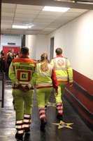Drei Mitglieder des Sanitätsdienstes in Leuchtwesten gehen in einem Gang. Sie tragen Sicherheitskleidung in Gelb und Rot und gehen in Richtung einer Veranstaltung. Der Boden ist mit Sternsymbolen dekoriert.
