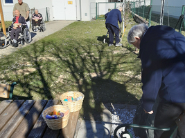 Eine Gruppe von älteren Menschen, einige im Rollstuhl, sitzt und steht im Freien. Eine Person beugt sich über den Rasen, während andere in der Nähe auf Holzbänken sitzen. Im Vordergrund liegen Körbe mit verschiedenen Gegenständen. Es ist ein sonniger Tag.