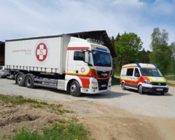 großer LKW Samariterbund Wien, daneben kleinerer Transporter auf Straße, daneben Feldweg