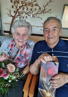 Ein älteres Paar sitzt lächelnd zusammen, umgeben von Blumen und Geschenken.