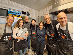 Sieben Personen stehen in einer Küche und lächeln in die Kamera. Sie tragen Kochschürzen mit der Aufschrift 