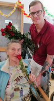 Eine ältere Frau hält eine rote Rose, begleitet von einem jungen Mann.